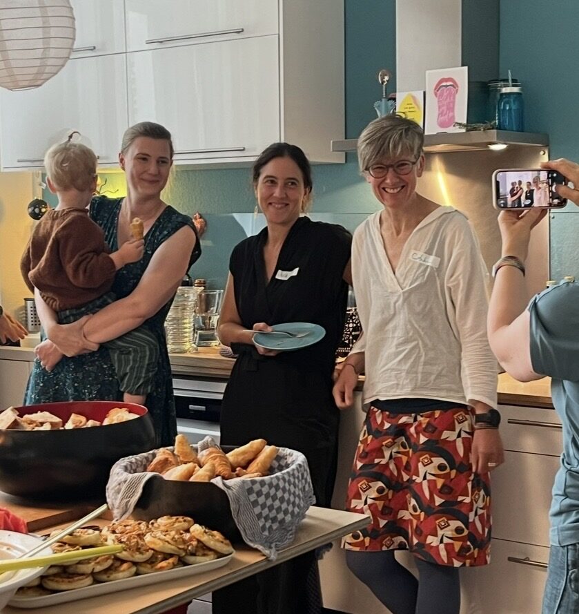 Vier Dozentinnen des montessori labors, eine davon mit Kleinkind auf dem Arm, stehen lachend an einem gedeckten Buffet in der Küche