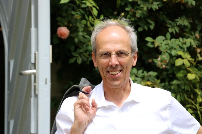 Thomas Helmle steht lachend im Türrahmen vor einem Rosenstrauch, ein Jackett über der Schulter